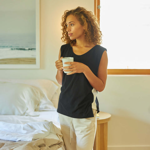 A young woman holding a cup of coffee while standing next to her bed and wearing a Slumber Cloud UltraCool Tank